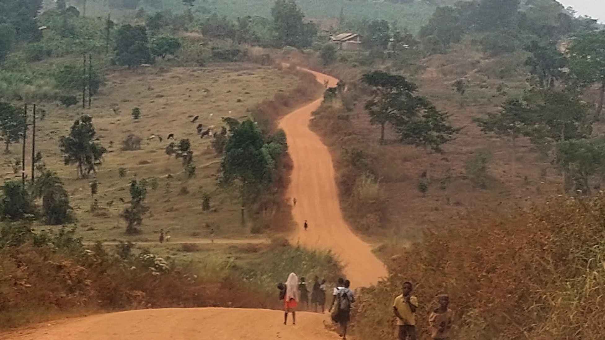 Dusty dirt road with trees around and people walking on the side of the road
