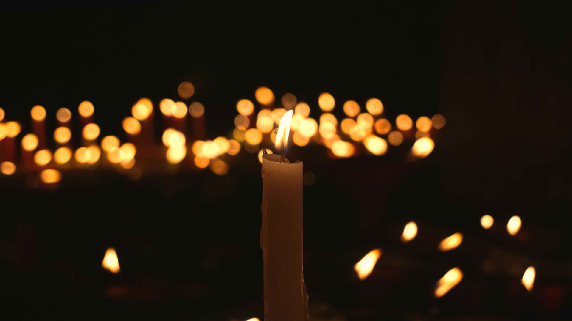Set against a dark background, a candle flickers in the foreground, surrounded by other candles.