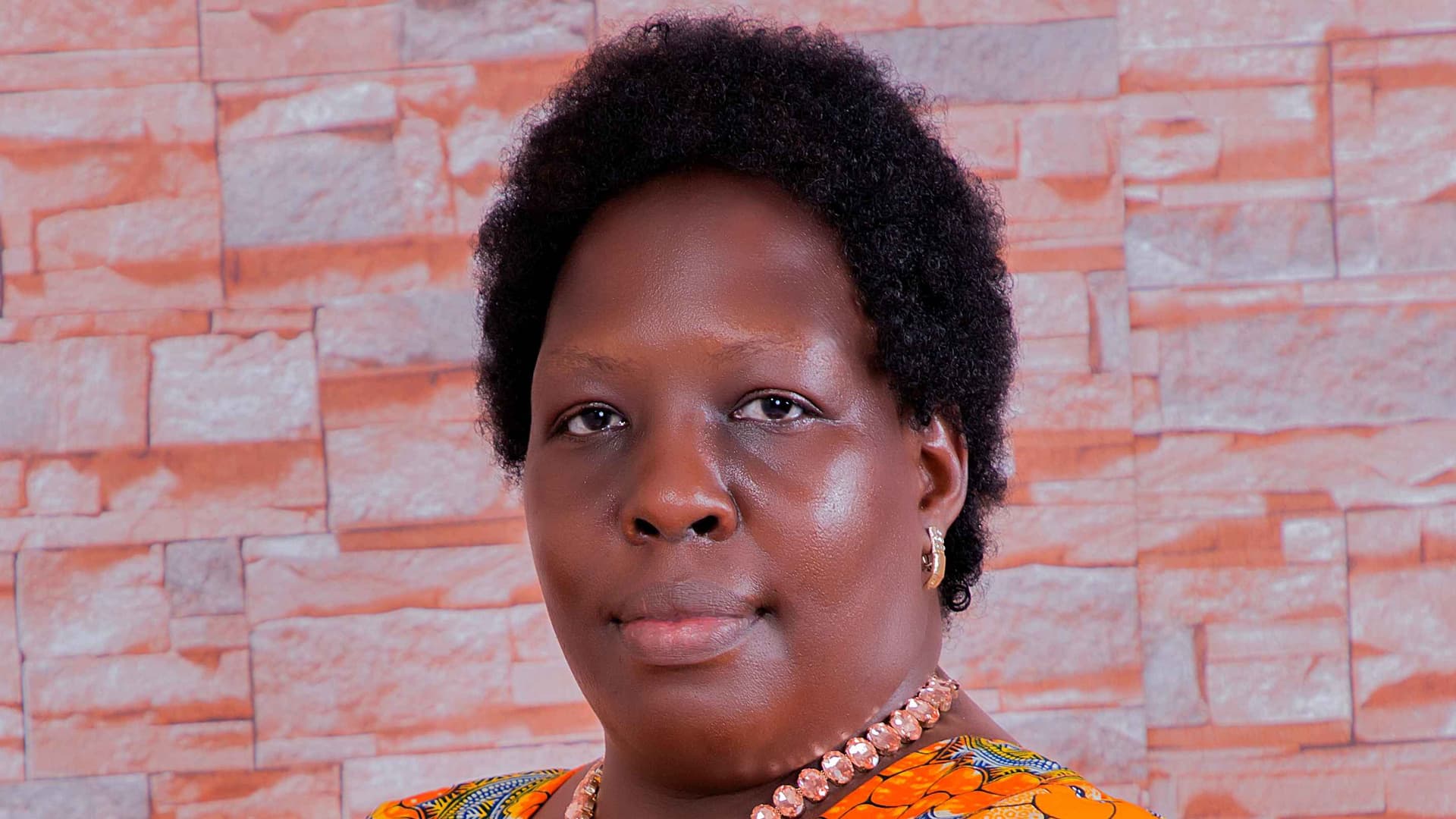 Ikirimat is a Black woman with short curly black hair. She is wearing gold earrings and a necklace with light-colored jewels and a bright orange-yellow printed top with hints of white and blue. She stands against a brick/stone wall.