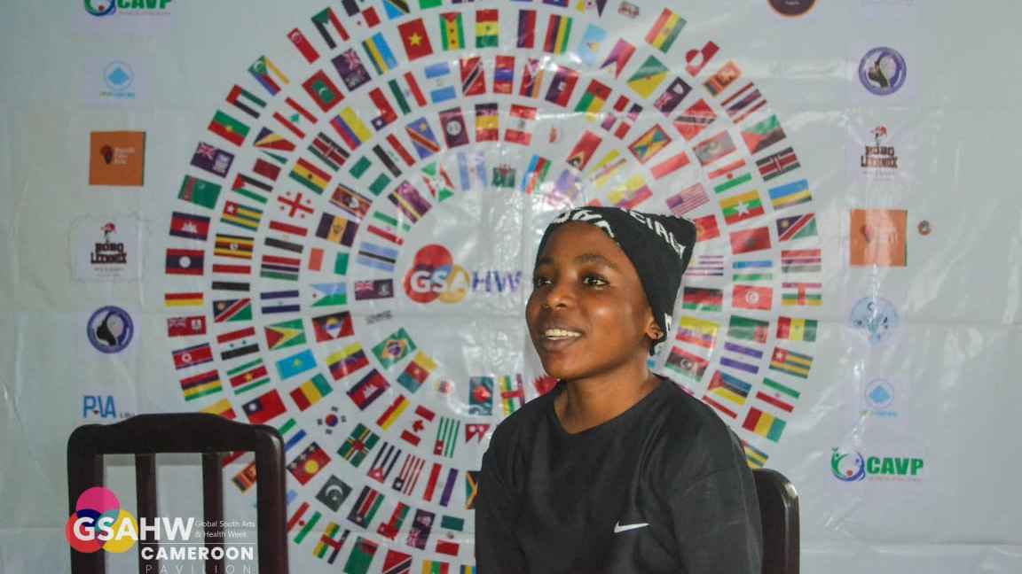 Young girl wearing a black hat and black sweater seating in front of a banner with maps of different countries organized in a circle. 
