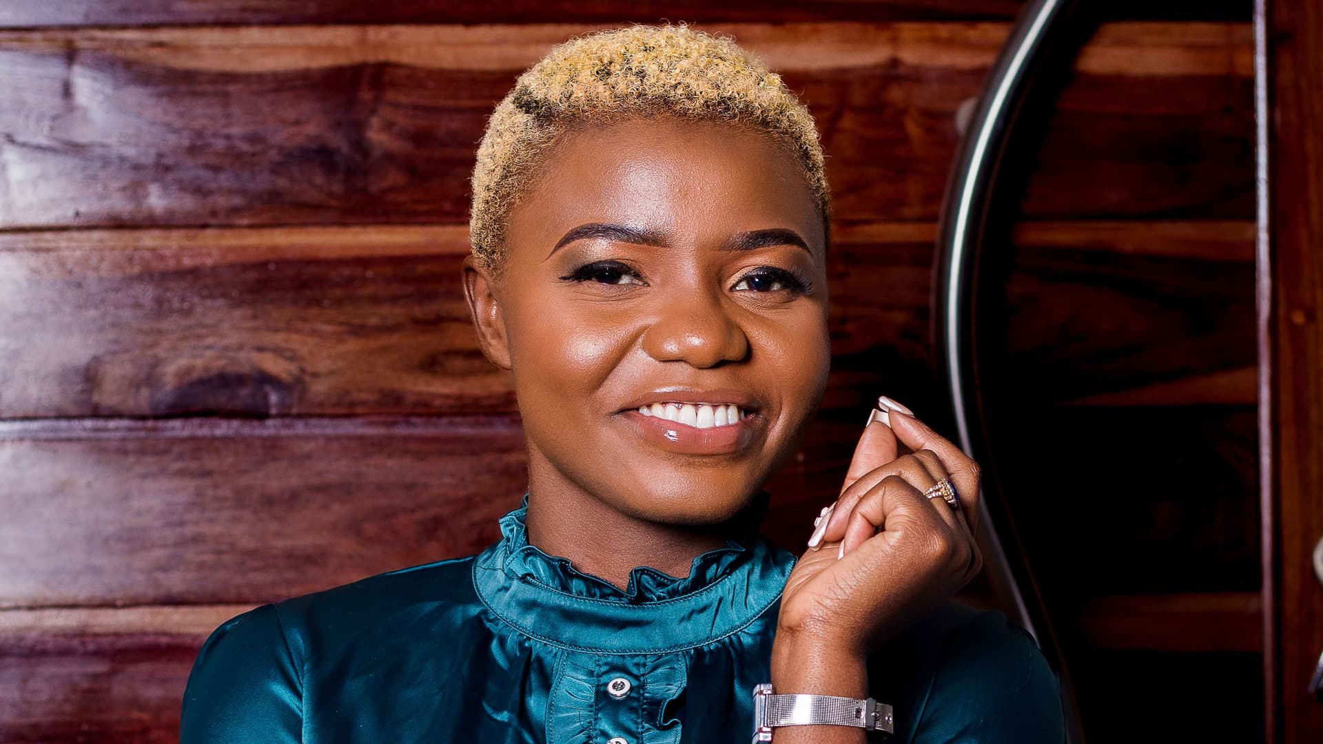 Sympathy Sibanda is photographed in a blue, long-sleeved blouse with ruffles and buttons. Her arm is pointed up with a silver watch. She has short blond hair and brown eyes and smiles in front of a wooden wall.