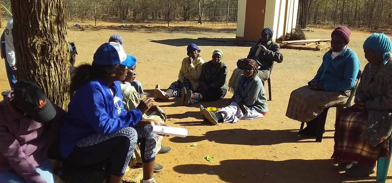 A community meeting taking place outdoors in a rural area, where a young female leader is sitting with local community members discussing issues and solutions together.