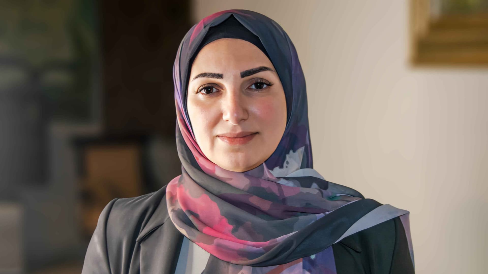 Hawraa Ghandour is photographed in a pink, purple, black, and gray headscarf over a dark colored top. She has tan skin and brown eyes and eye brows. The background is dark on the leftside and light on the right. 