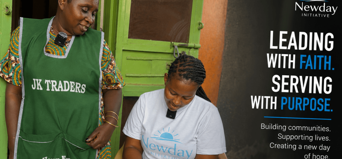 A young woman wearing a New Day Initiative T-shirt sits beside a community member while taking notes during an outreach visit. The visit focuses on listening to families and understanding their needs.