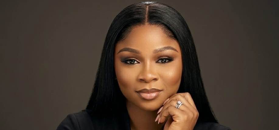 Professional headshot of a Nigerian woman with long dark hair, wearing a black outfit and resting her hand gently under her chin. She is looking directly at the camera with a calm and confident expression against a dark background.