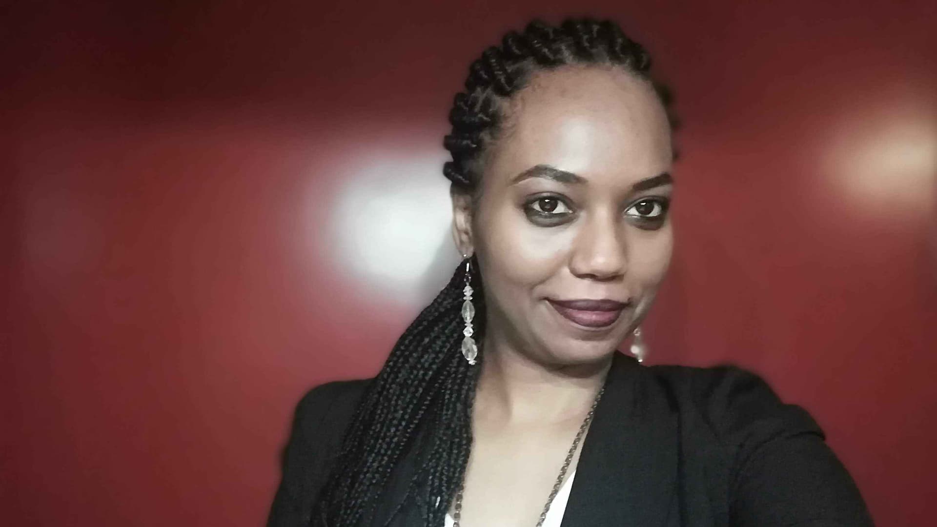 Amina is photographed with long, dark braided hair, a white blouse, and a black blazer. She wears beaded earrings and a chain necklace. She smiles in front of a red backdrop.