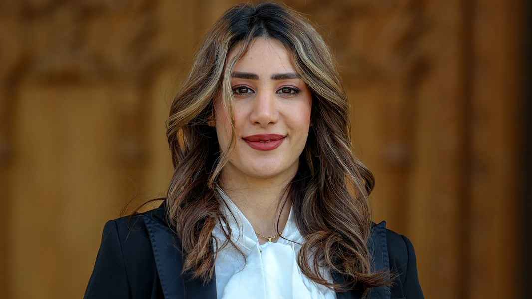 A professional portrait of a woman with long brown hair wearing formal attire, photographed indoors against a neutral background.