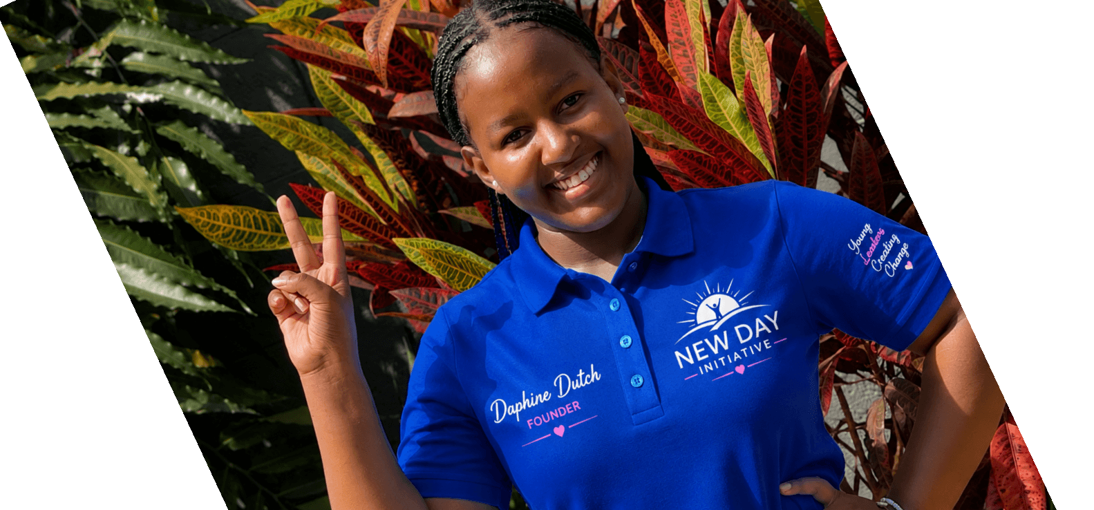 A smiling young woman wearing a blue New Day Initiative founder shirt stands outdoors in front of colorful plants and holds up a peace sign.