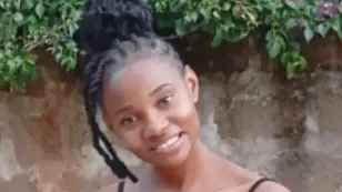 Faith has black braided hair that is pulled up and is smiling at the camera in front of greenery hanging over a wall.