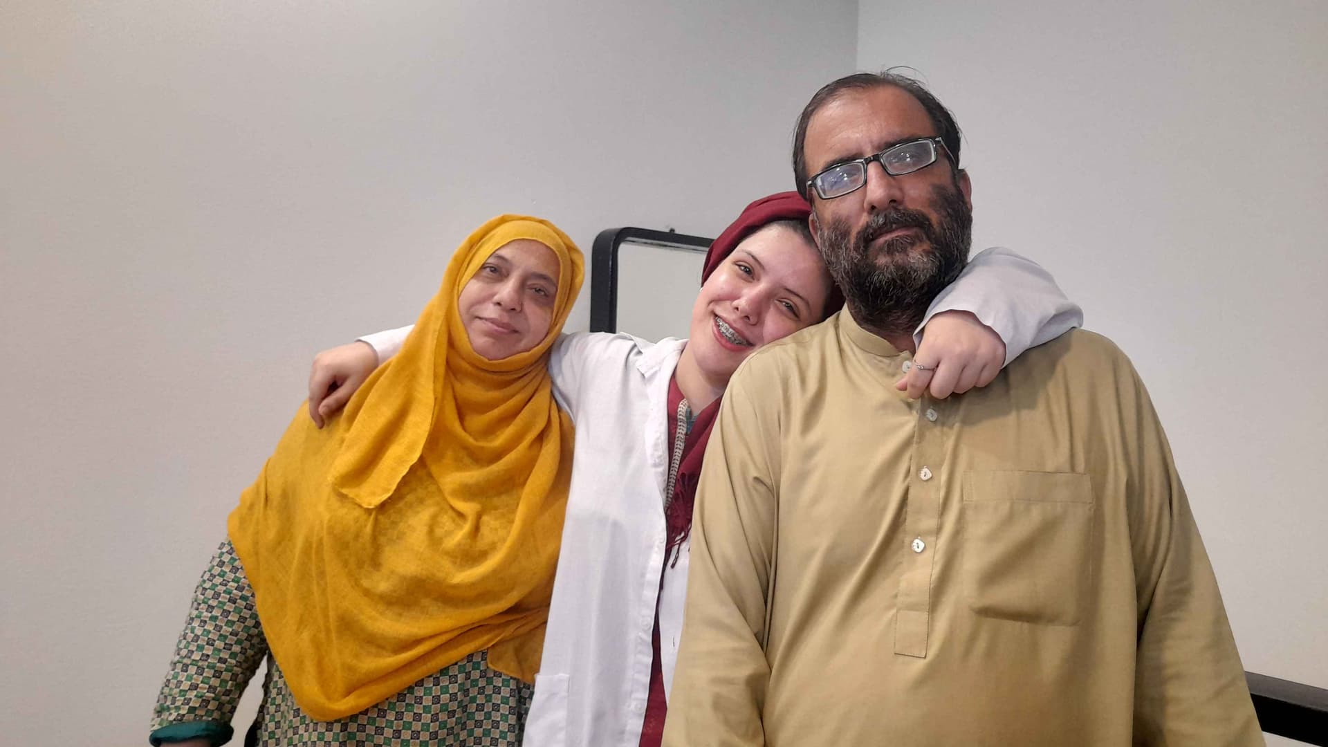 Author Iqra Jan, center smiles in a red head covering and white blouse. Her mother, left wears a checkered top and yellow head covering. Her father, right, wears a beige button-up shirt.