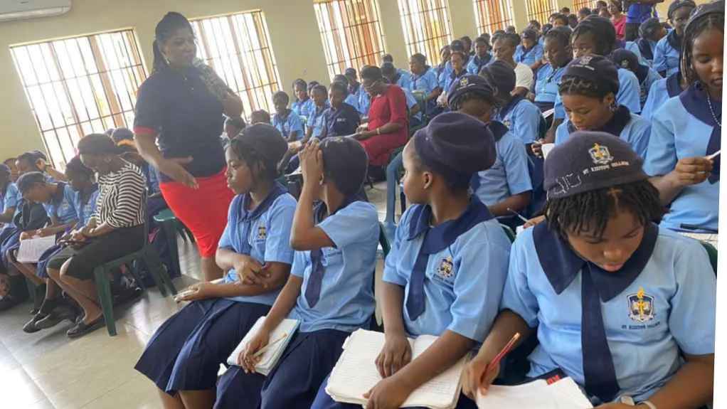 Ruth Okoirhon, a woman in a navy blue shirt and red skirt, speaks before a classroom full of girls in uniforms of light blue shirts and navy blue skirts. The girls are seated in rows of chairs.