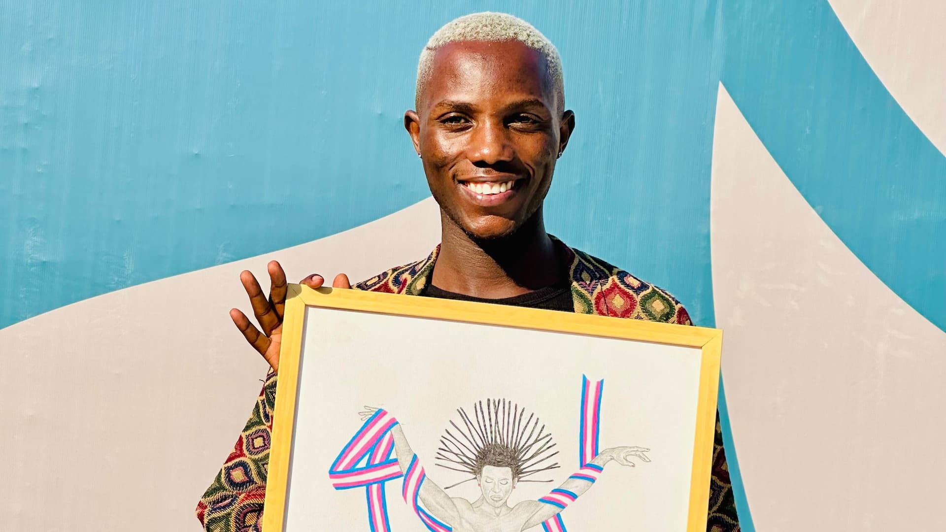 Myself smiling while holding a framed piece of colorful artwork, standing against a blue background.