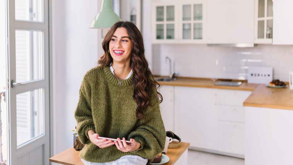 A smiling woman in a green sweater sits on a kitchen counter, holding a phone in a bright, modern kitchen. T
