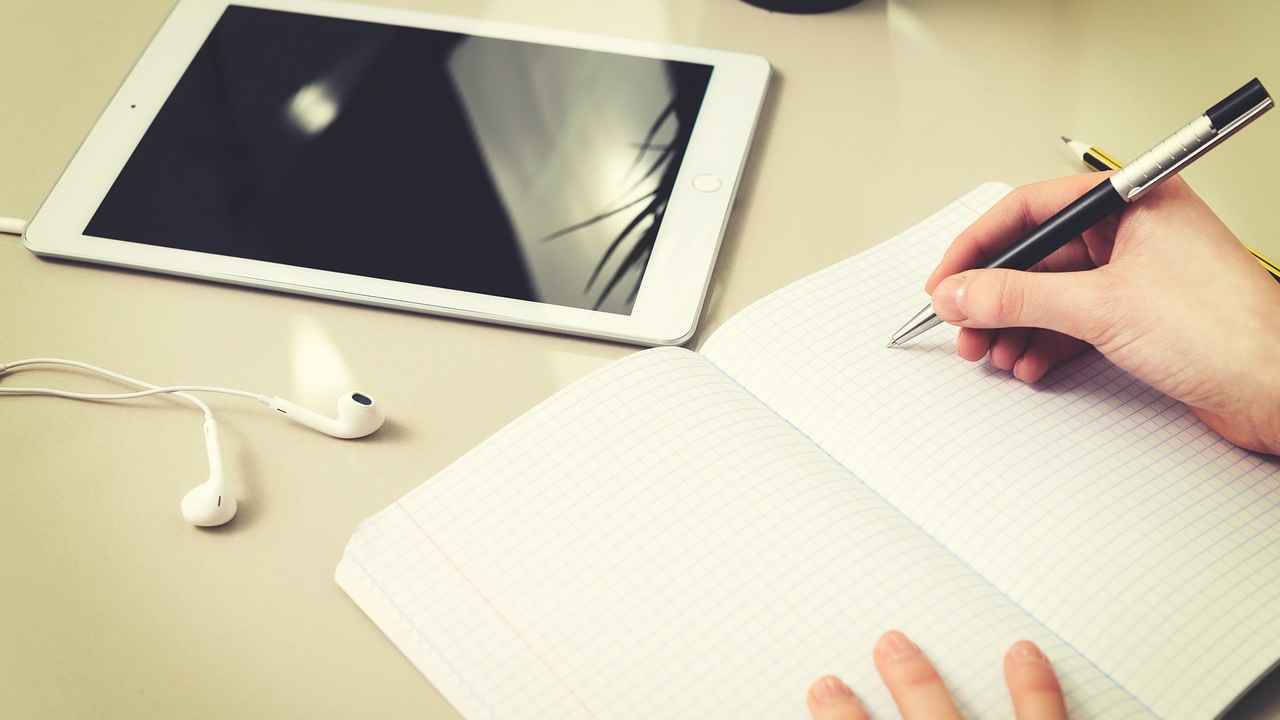 A hand holding a pen over an open notebook, with a smart tablet and earphones placed nearby to suggest that a person is learning