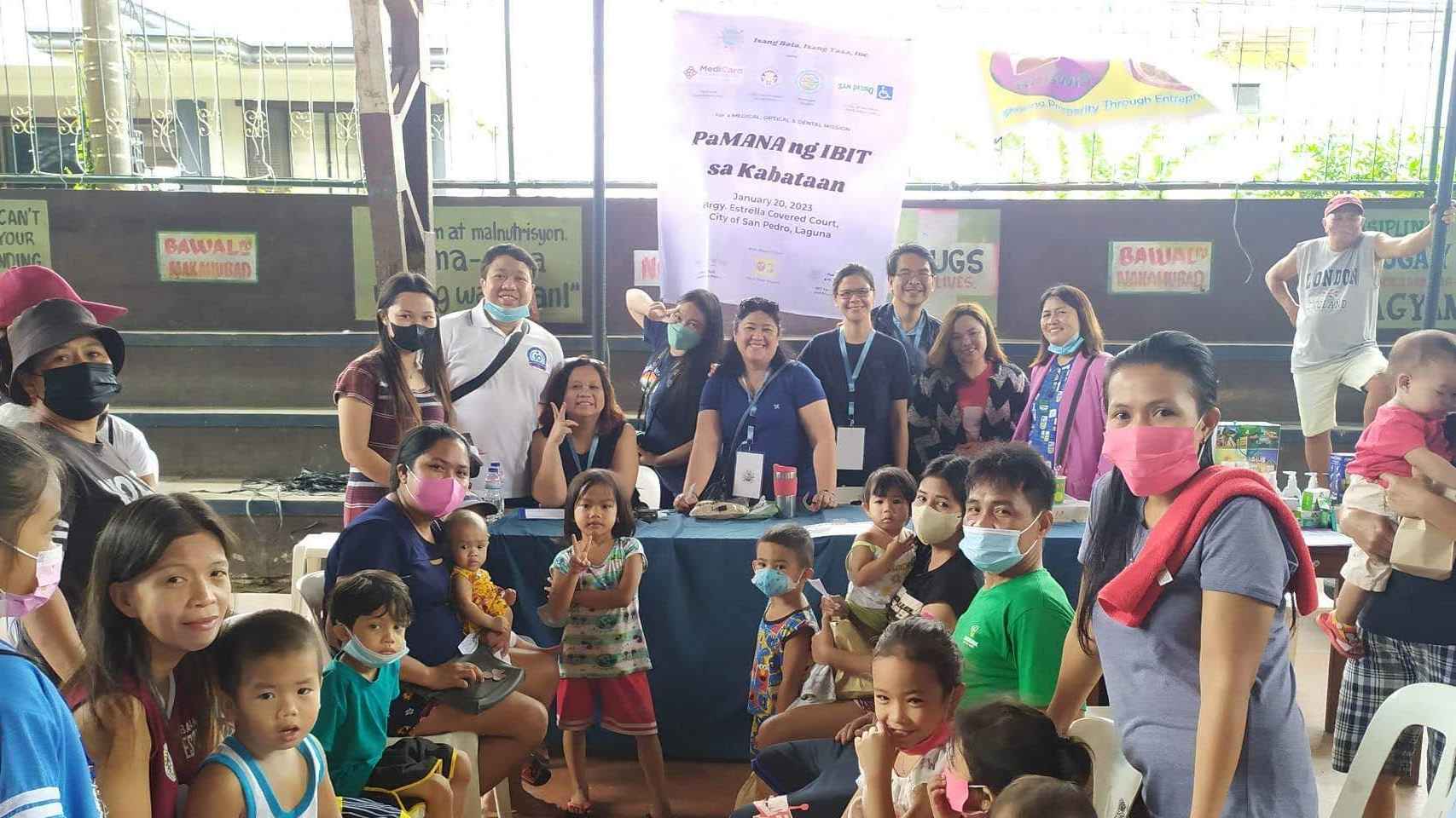 In an open space with banner in the background, and several people in blue standing in front of the banner smiling together with the children and parents registering to receive the hygiene and food packs from IBIT 