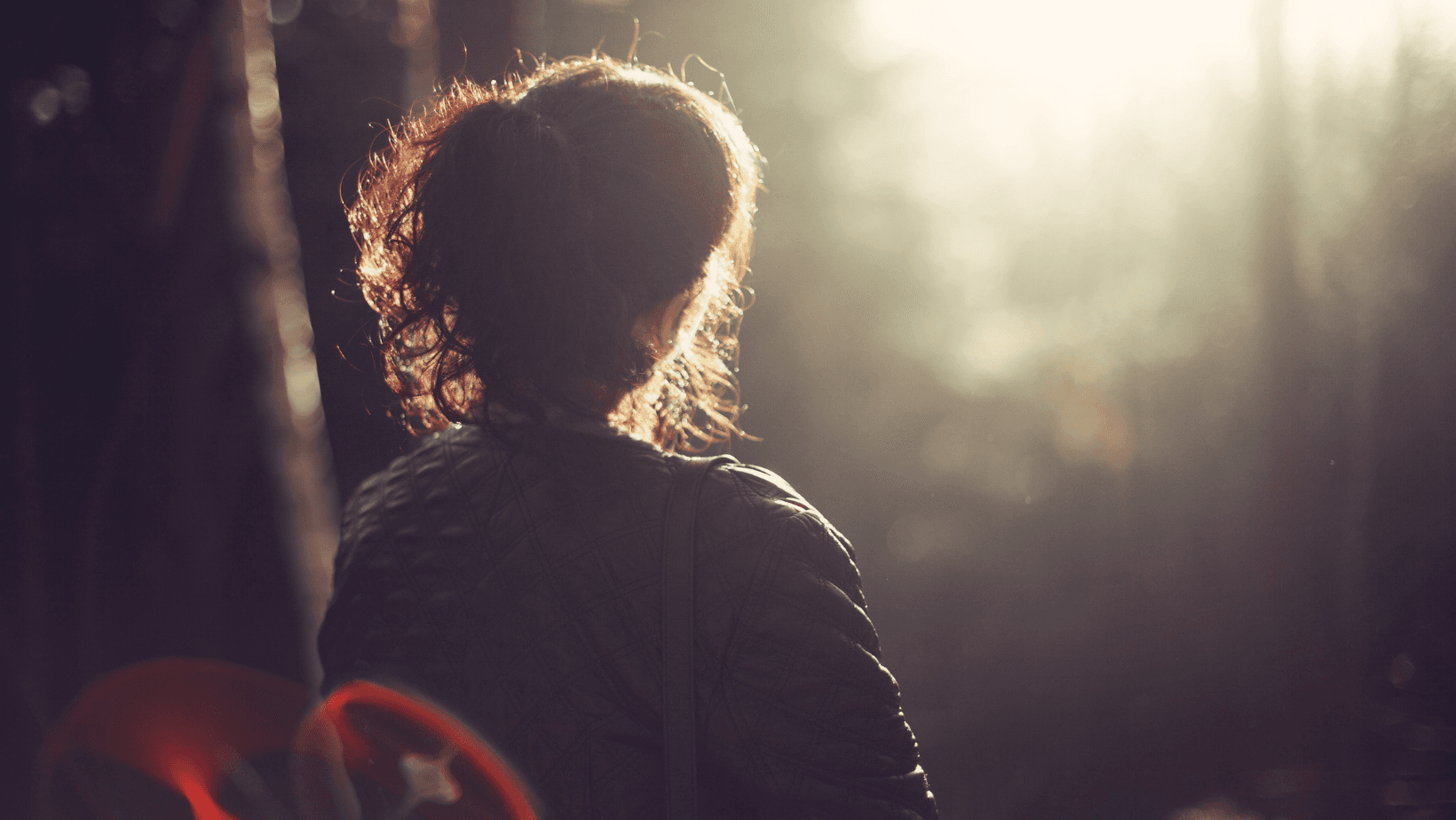 A woman looks toward the sun. She wears a ponytail and jacket and appears to be in a forest setting.