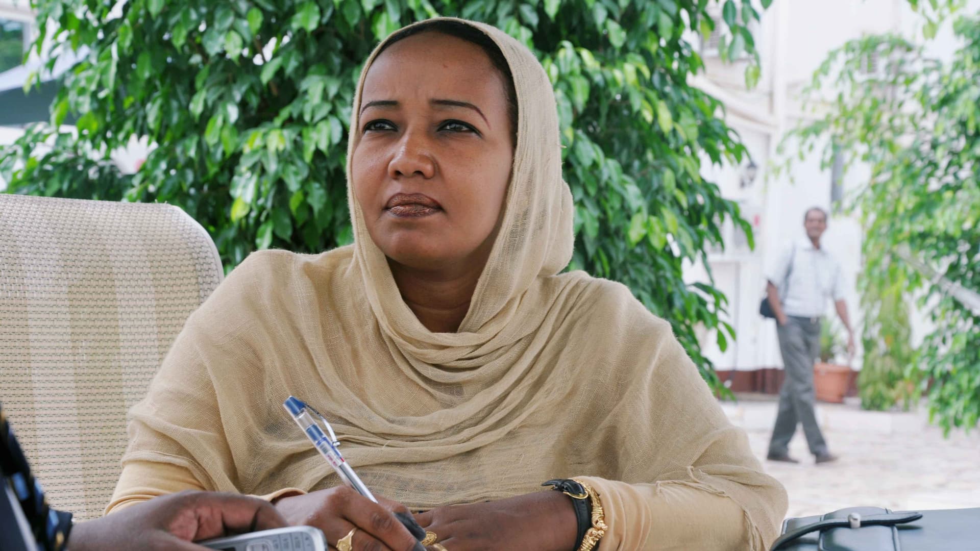 Nadra AlMahdi is photographed wearing a beige long-sleeved top and head scarf. She has brown skin and black hair and a black watch and gold jewelry. She sits in a chair with a pen and paper. Behind her is greenery and a man walking along a patio.