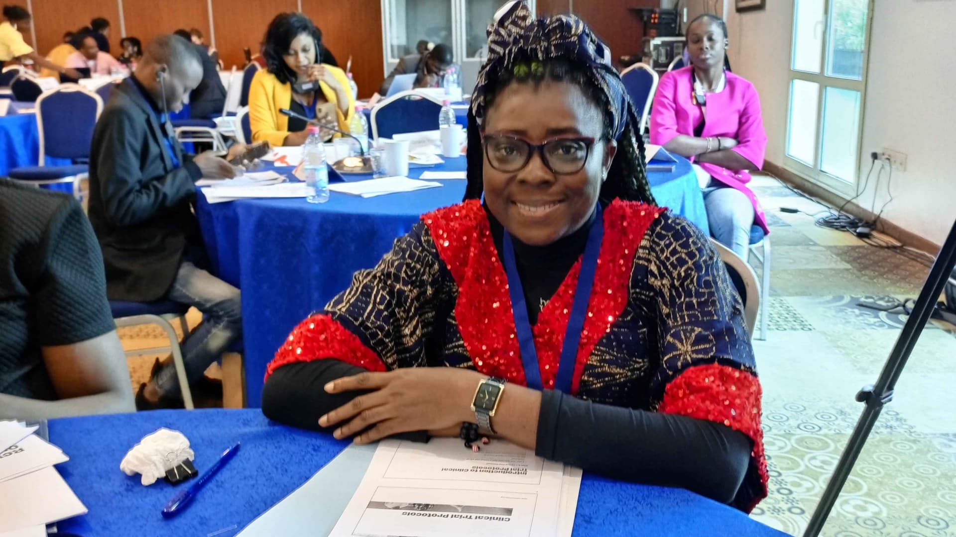 I am attending a training conference here., sitting with my hands on the table, wearing a dark dress with red accessories with a headtie to accentuate my look, a red and black coloured glasses and a beautiful smile on my face.