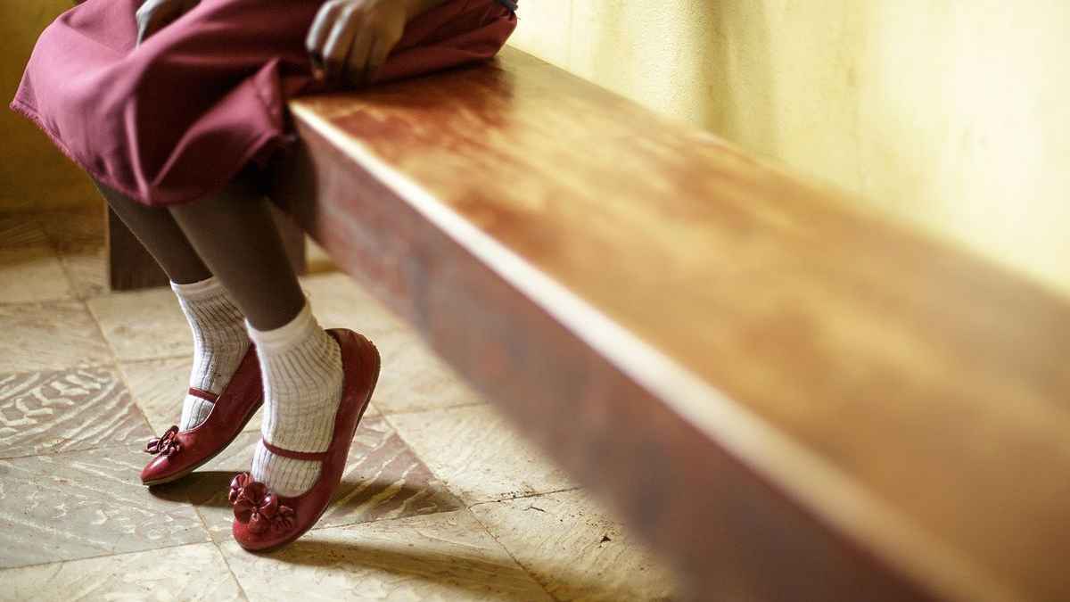 A child sitting on a wooden bench, wearing a maroon skirt, white socks, and red shoes with bows. The child's hands are resting on the edge of the bench, and their legs are hanging down. The background includes a yellow curtain and a tiled floor.