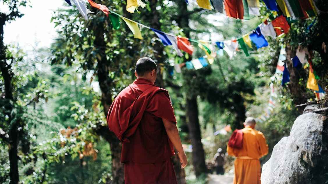 A man in a red robe and a man in a yellow robe walk under strings of prayer flags. The are surrounded by lush green scenery.
