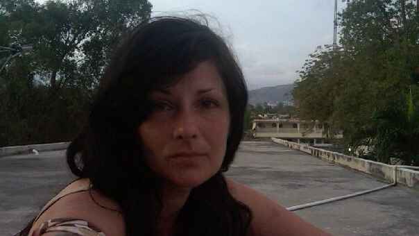 Amanda, a woman with brown hair and eyes, looks into the camera during a visit to Haiti in 2010. Behind her is a parking lot and greenery.