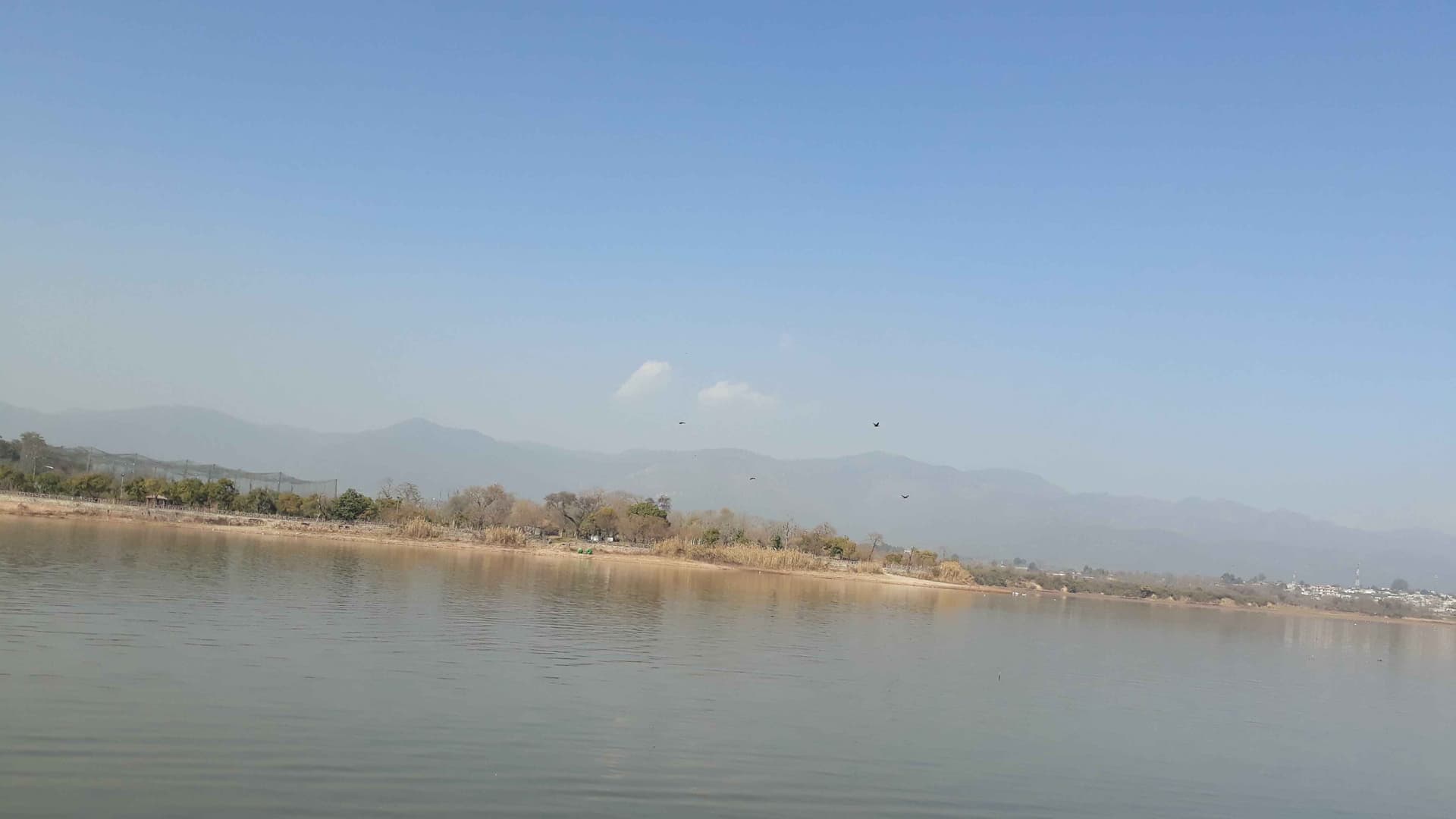 This scenic photo features a body of water in Pakistan surrounded by sandy banks, trees, and mountains.