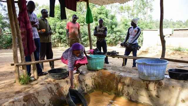 A woman wearing pink and purple fetches water with a black bucket. Surrounding her are five men who look on under and awning.