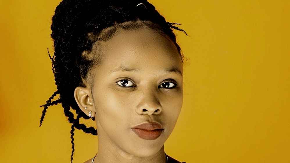 Purity Waithira, a woman with black pulled back hair and brown eyes, looks into the camera. Behind her is a light orange background.
