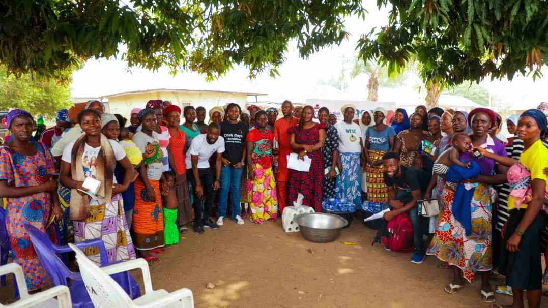 A large group of women dressed in colorful skirts and headscarves gather under a tree in Buzunkure Village.